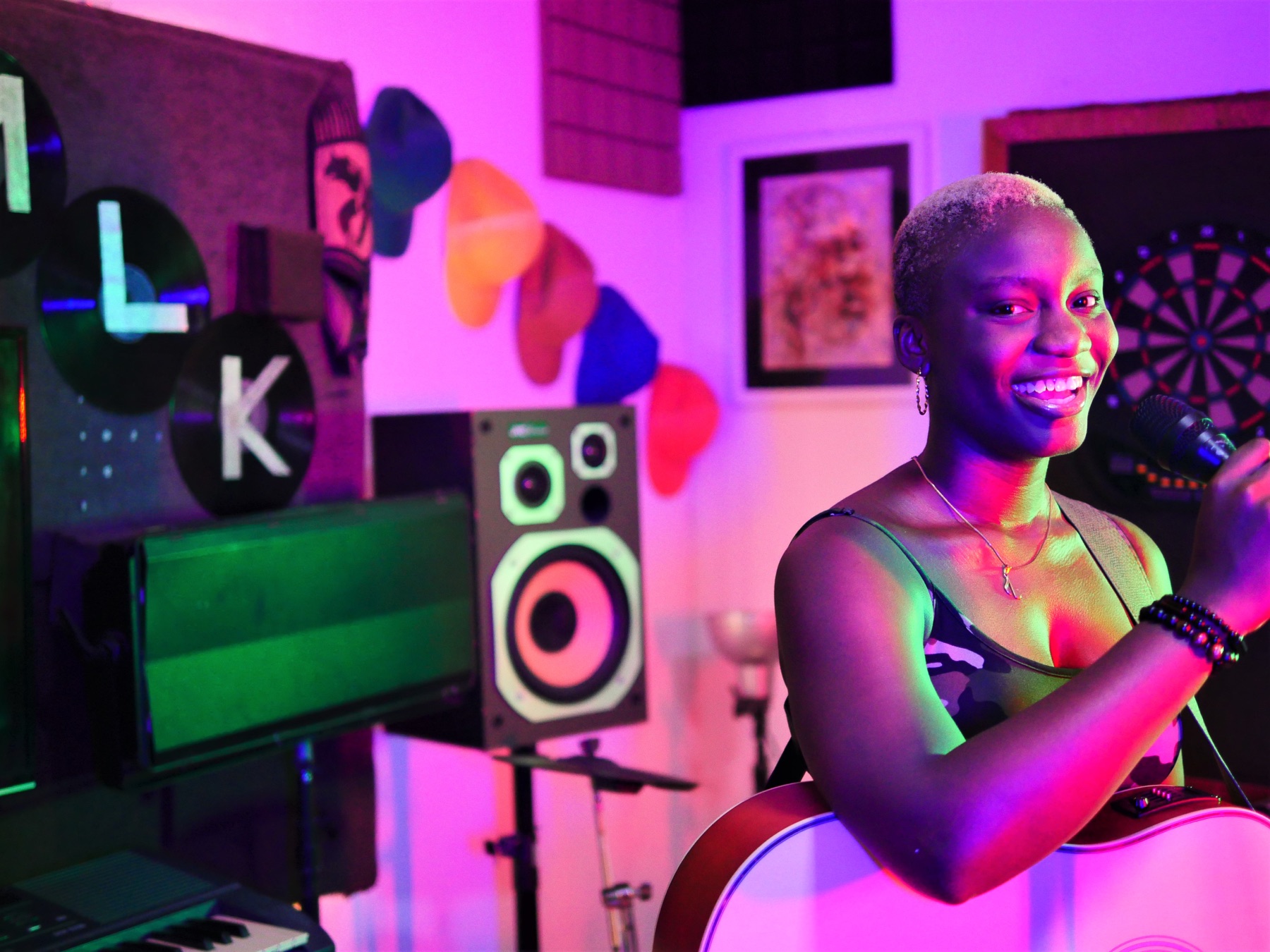 KKC smiling with a microphone and guitar in a colourful studio setting.